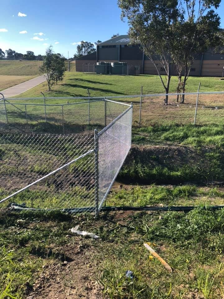 Broken chain-link fence beside a grassy roadside lot and a building under a sunny sky