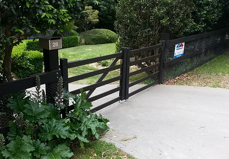 Black wooden gate across a driveway beside a green hedge and garden path