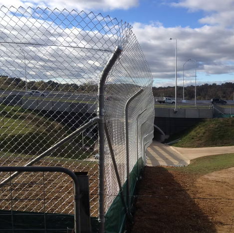 Chain-link fence along a roadside path under a cloudy sky, with a road and bridge in the background