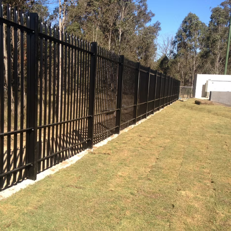 Black metal fence along a grassy yard beside trees and a white building on a sunny day
