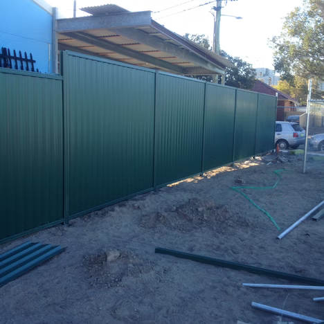 Green corrugated metal fence beside a dirt construction area with scattered pipes and a car in the background