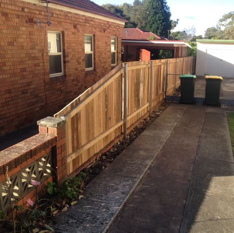 Brick house side path with a new wooden fence along a driveway and two green bins at the end.