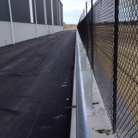 Narrow asphalt walkway between a building and chain-link fence, with a metal guardrail along the edge.