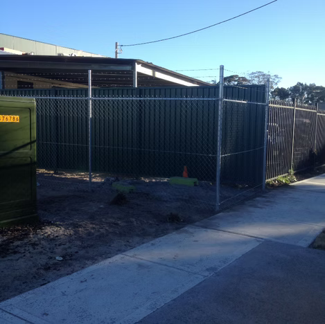 Chain-link fence beside a sidewalk and driveway, with a covered yard and industrial building behind it.