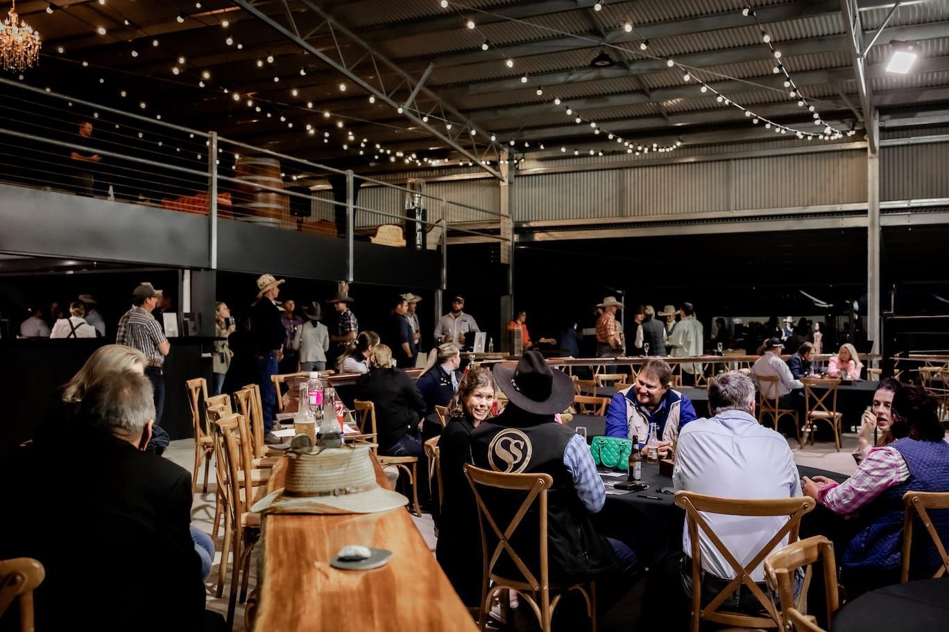 People at tables in an event space, some wearing hats. String lights overhead.