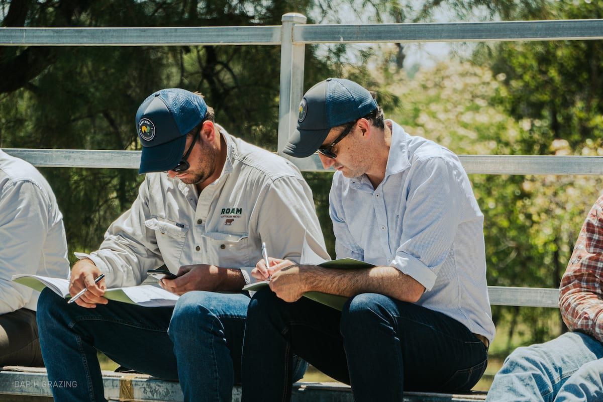 Two men in hats, outdoors, seated and reviewing documents; fence in background.