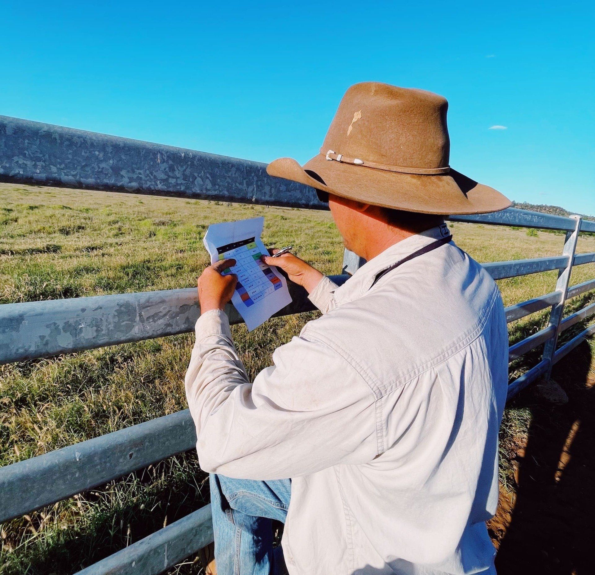 Man in a hat leaning on a fence, looking at a document outdoors in a field.