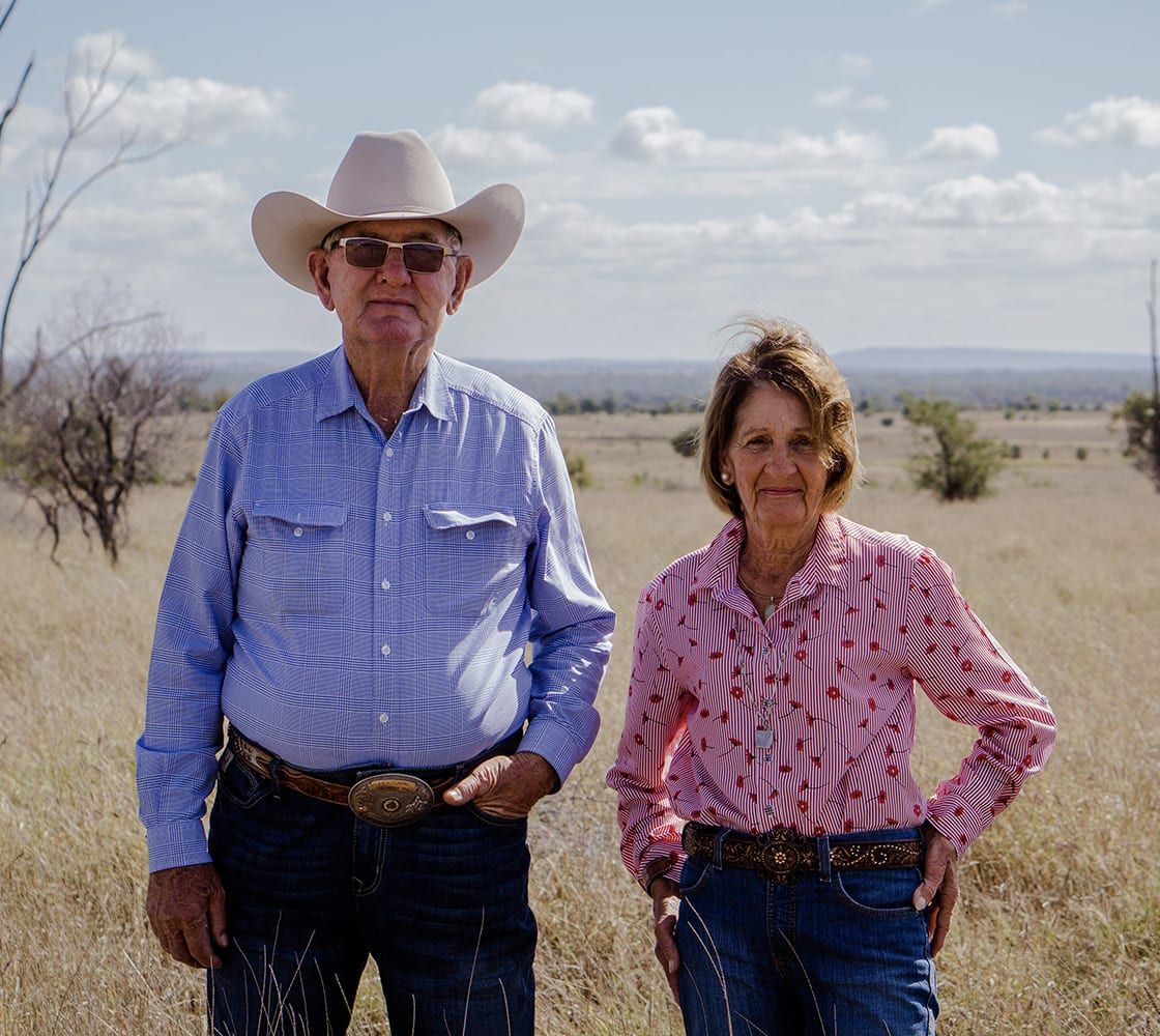 Elderly couple in cowboy attire stands in a grassy field under a blue sky.