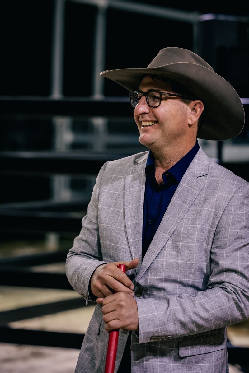 Man in cowboy hat, sunglasses, and work shirt smiles. Leans against fence, field in background.