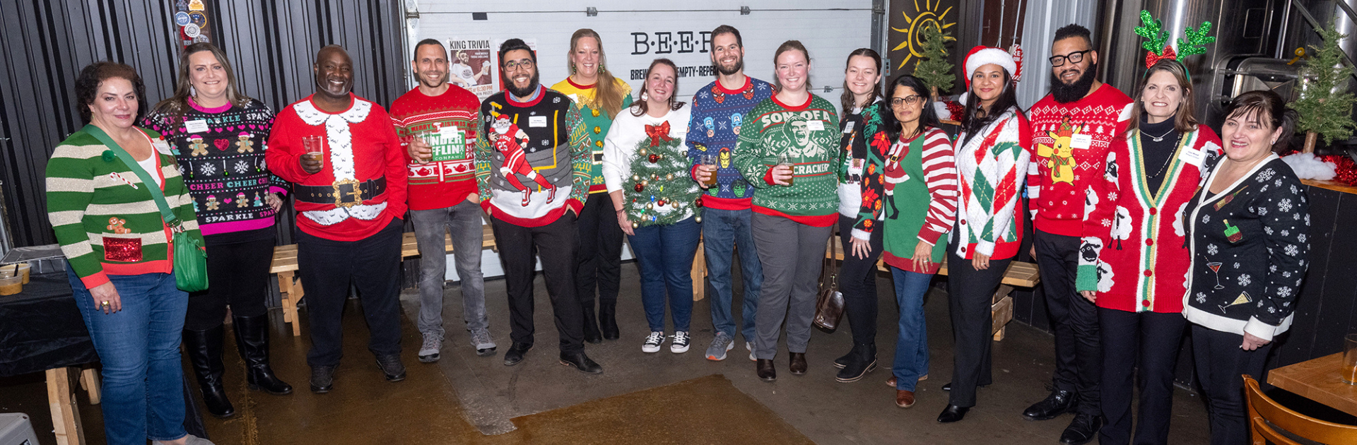 Group of people wearing festive Christmas sweaters posing indoors. Several have holiday-themed accessories.