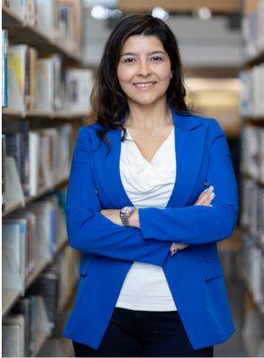 A woman in a blue jacket is standing in a library with her arms crossed.