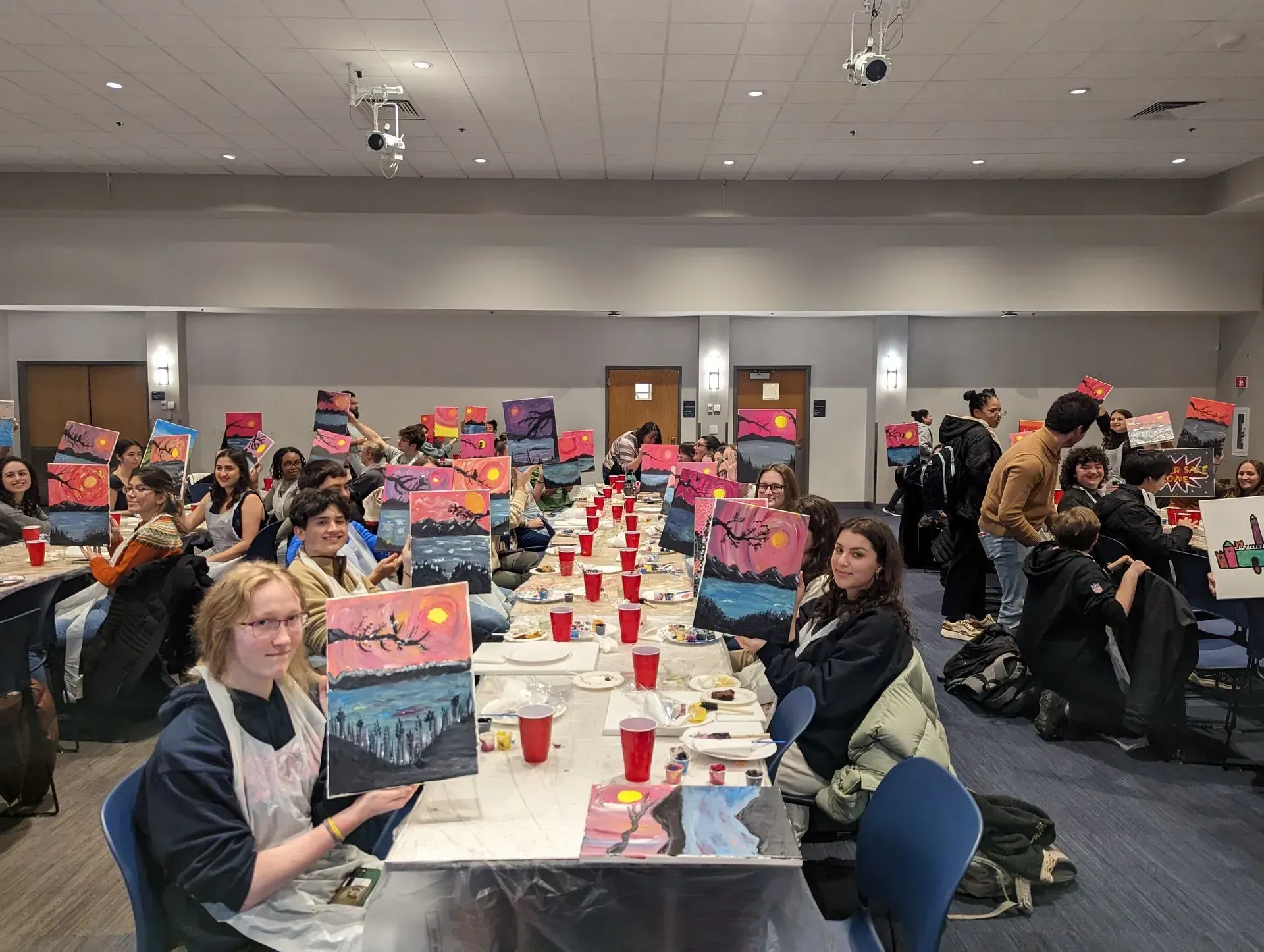 Group of people at a paint and sip event, holding up their landscape paintings. They're in a large room with long tables.