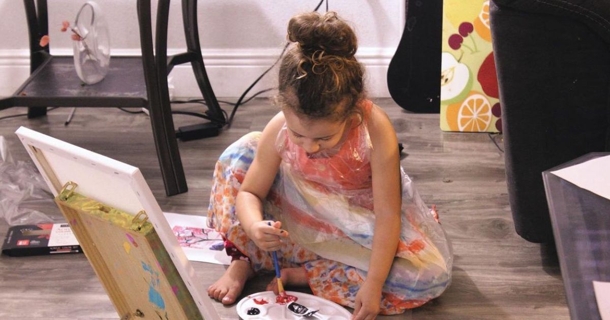 A little girl is sitting on the floor painting on a canvas.
