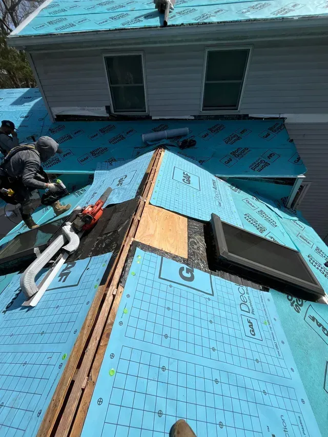 Roofers working on a roof with blue underlayment. One roofer is using a nail gun. The setting is outdoors on a sunny day.