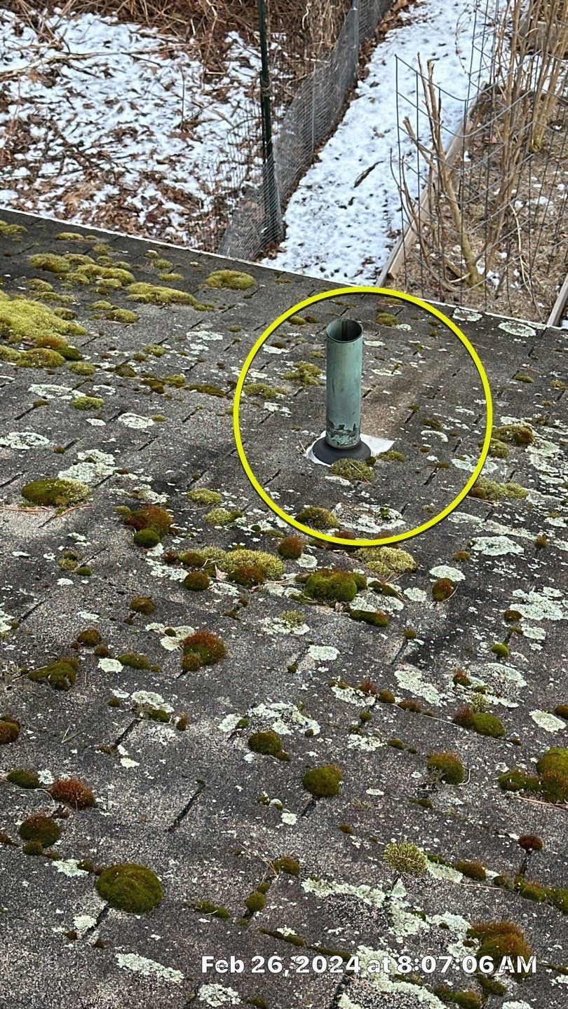 A close-up of a weathered rooftop with green moss, a metal vent pipe circled in yellow, and snow in the background.