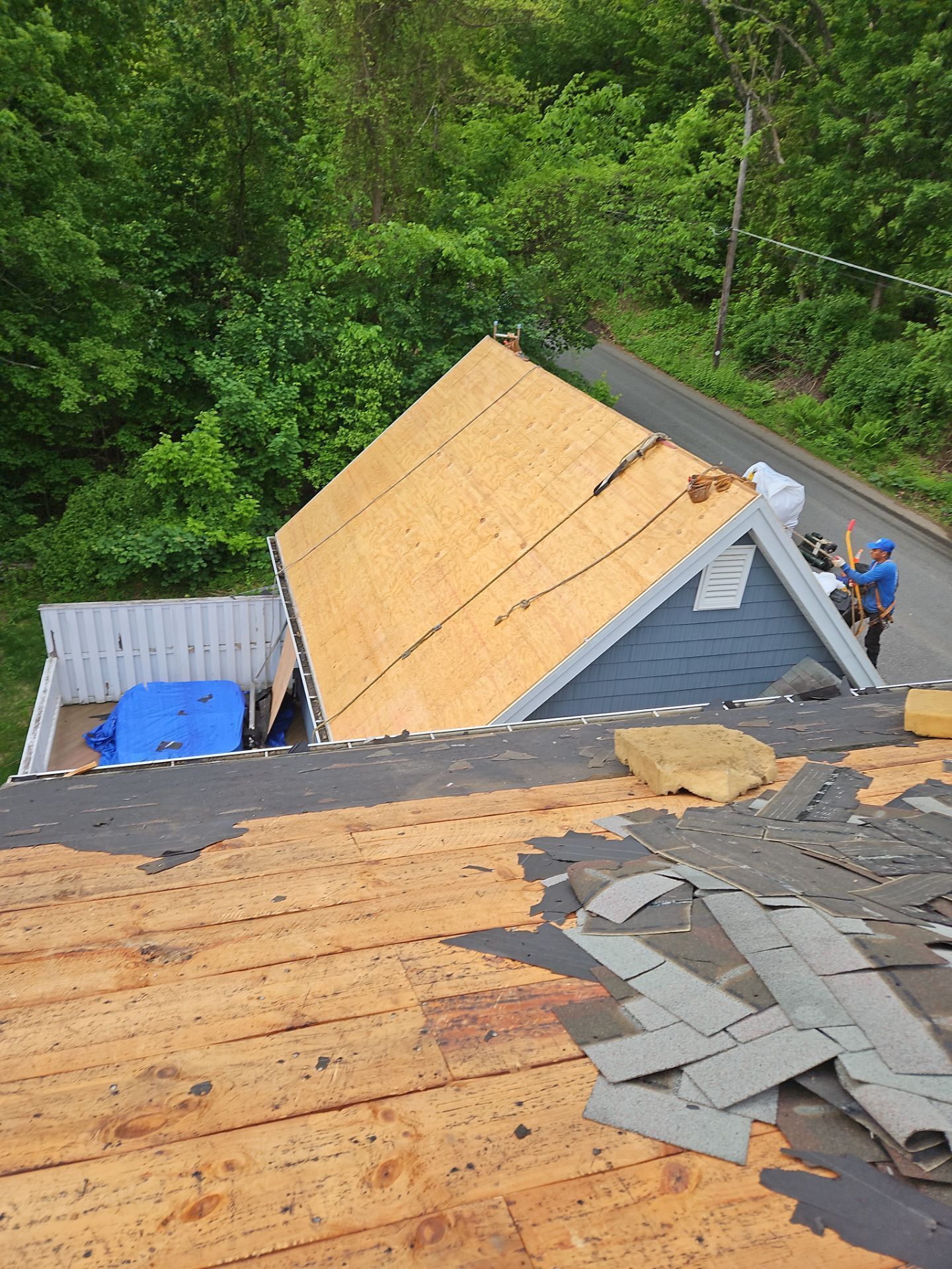 A partially re-roofed house with a worker, a blue tarp, and green trees. The old shingles are removed and new wood is exposed.