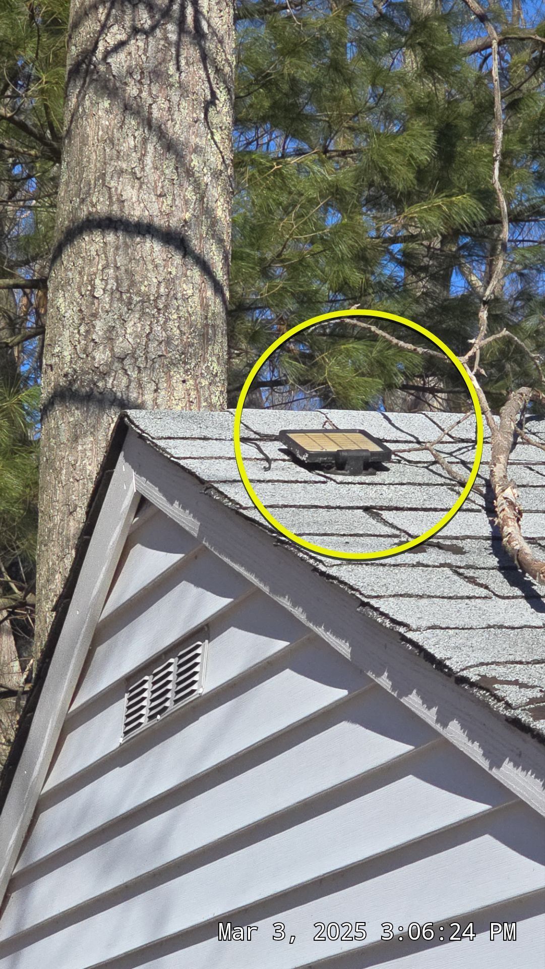 A rectangular roof vent circled in yellow on a gray shingle roof, next to a white house with gray siding and a tree.