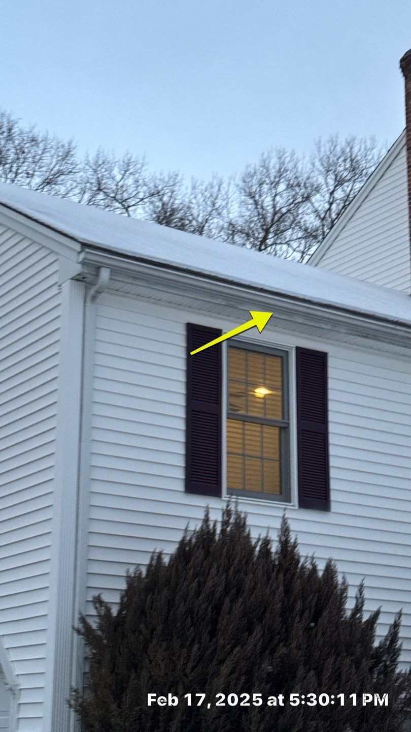 Snow-covered roofline of a white house with a window and black shutters; arrow points to the roof's edge.