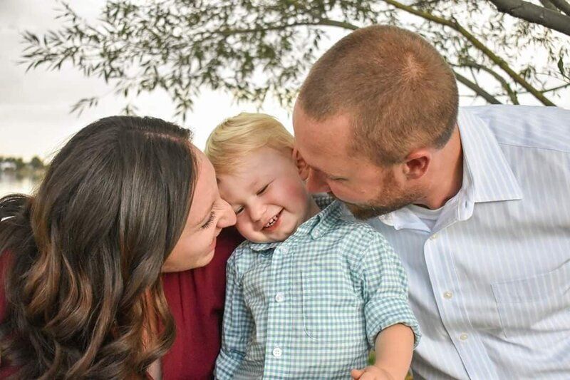 A dad and a mom are snuggling their toddler who is smiling between them. Family.
