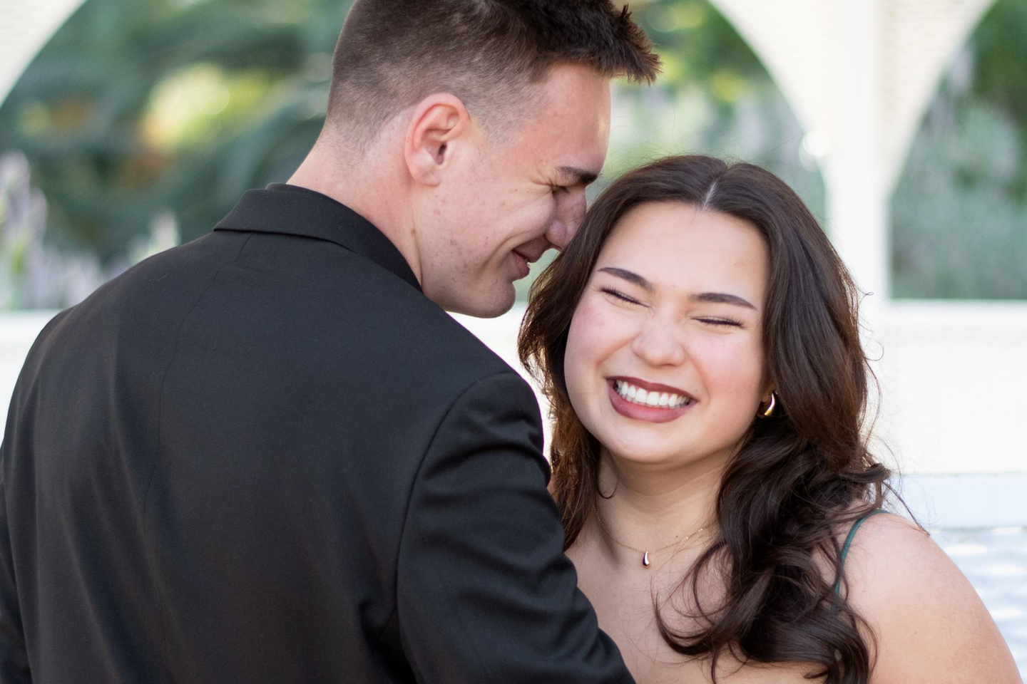 Man leans toward smiling woman, both are outdoors. He wears a black blazer, she has long dark hair and a necklace.