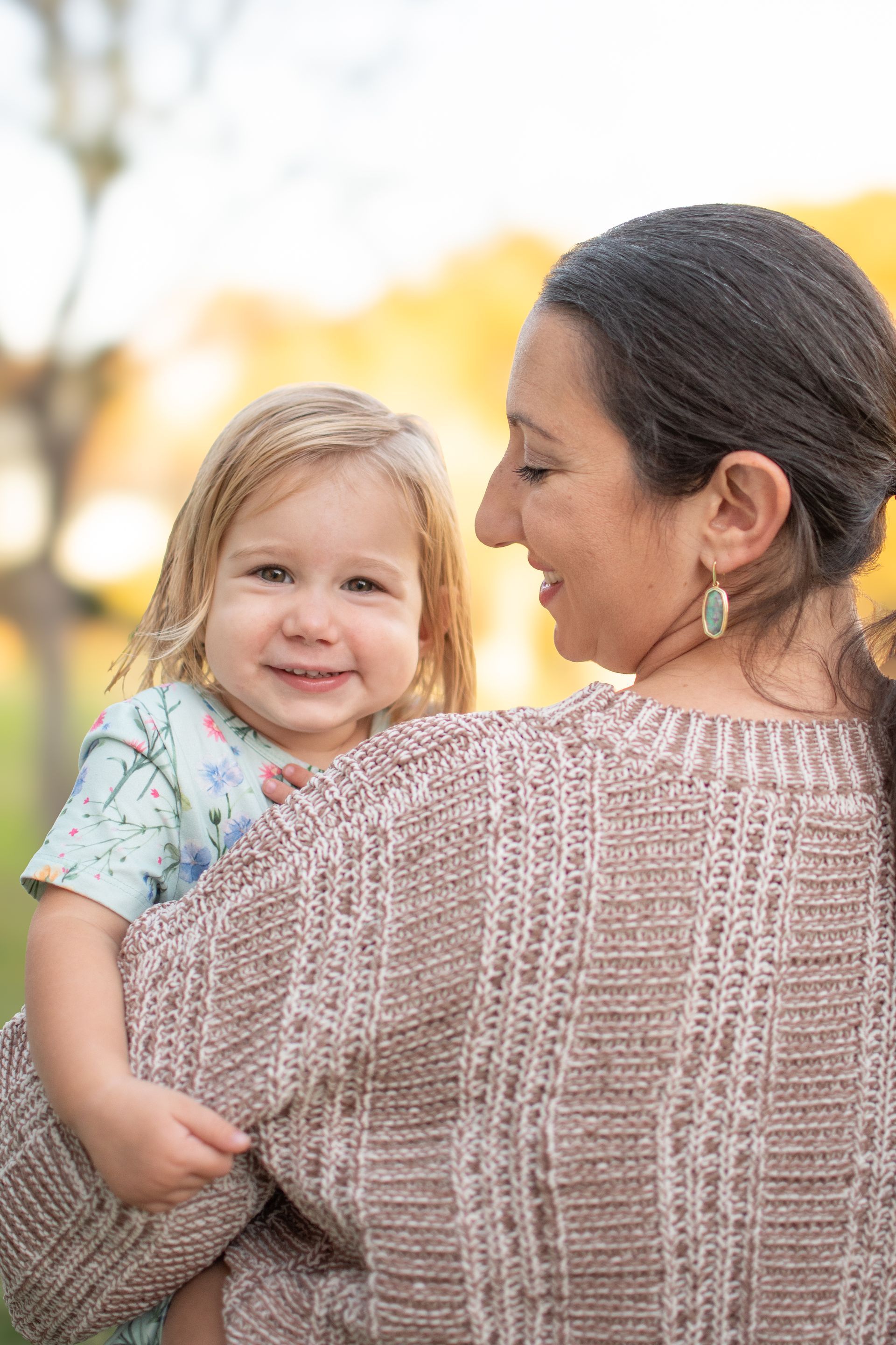 A woman in a textured tan sweater holds a smiling toddler in a patterned light blue dress 