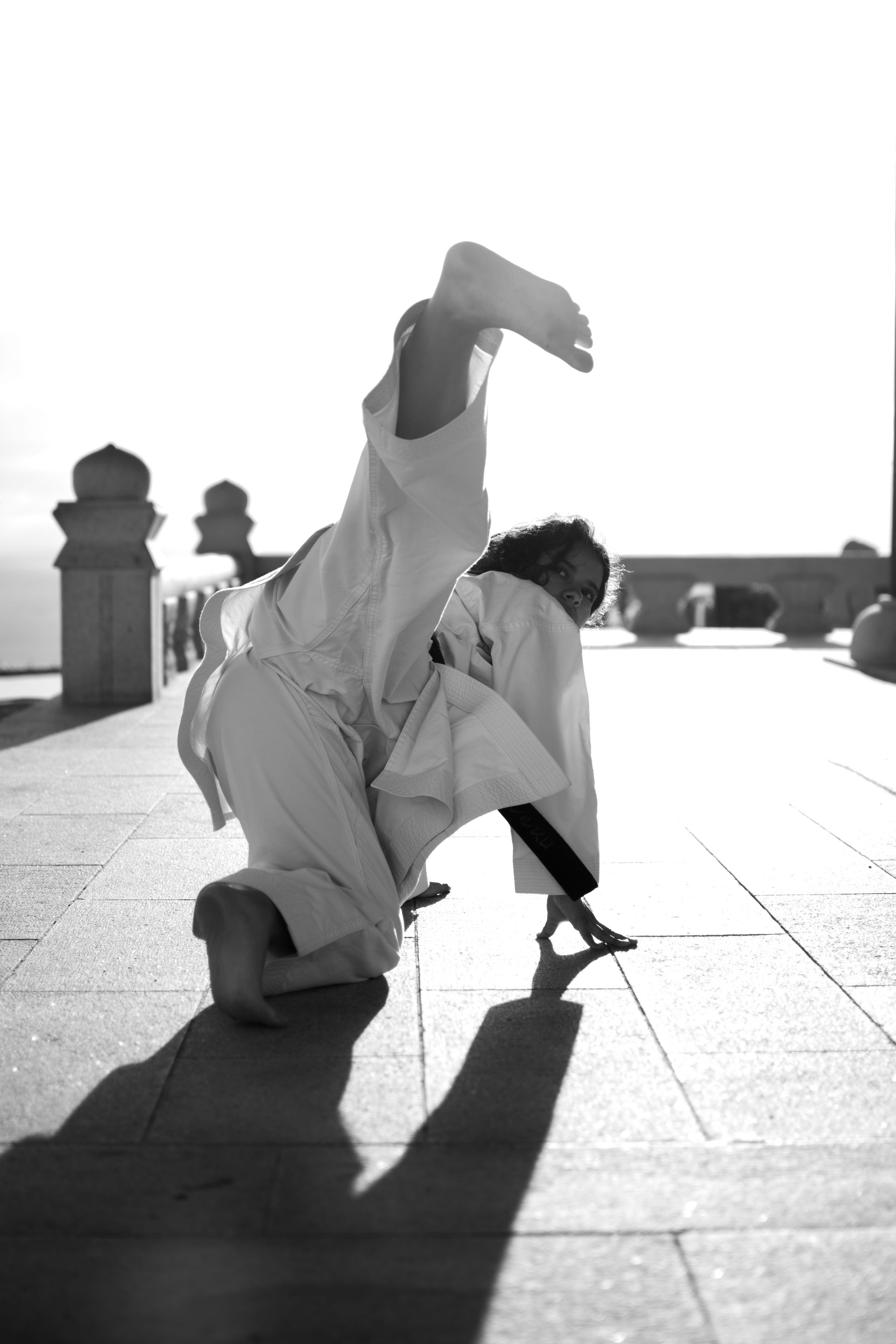 A person in a karate uniform performing a high kick on a stone patio during a bright, sunny day.