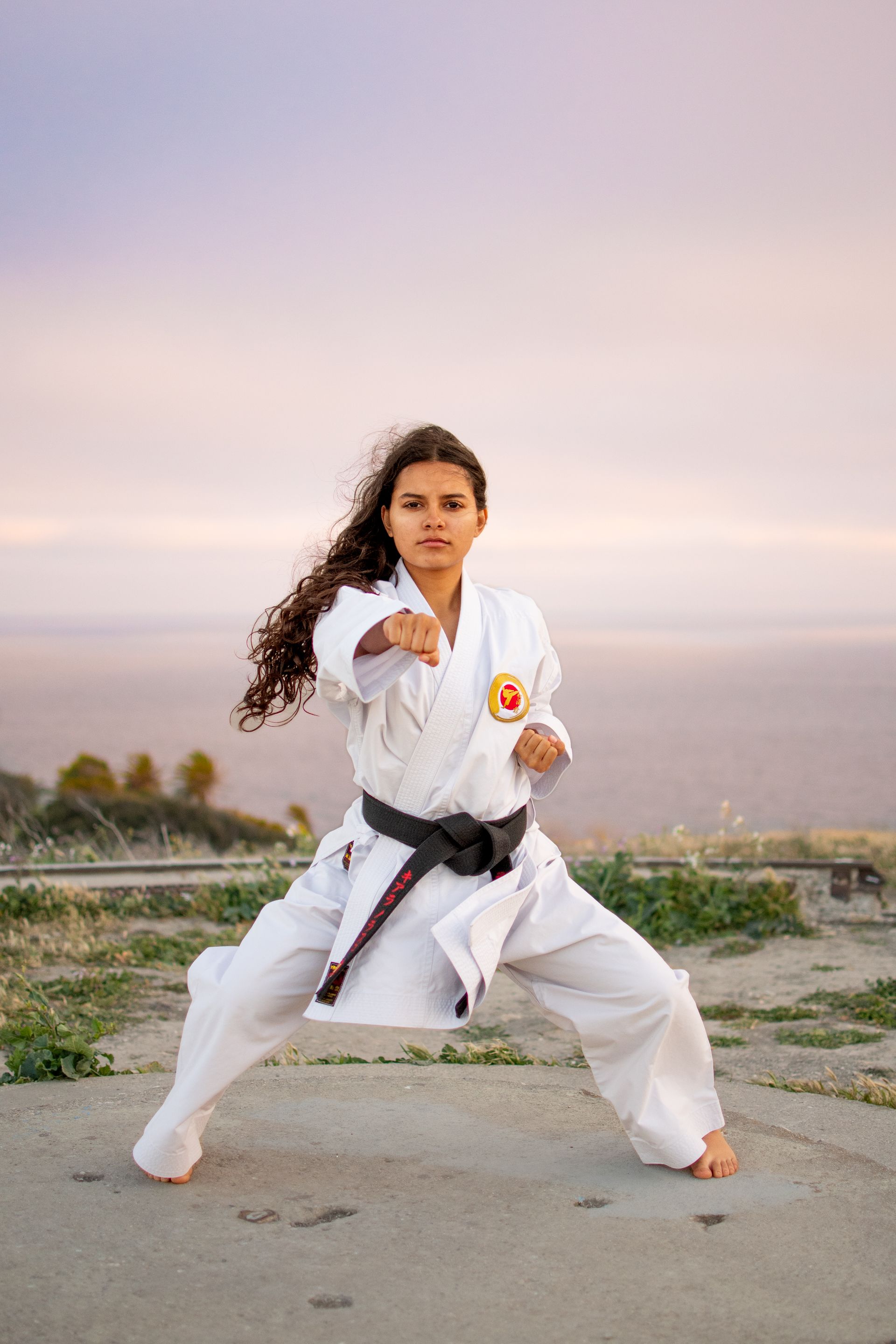A person in a white karate uniform and black belt performs a punch in a wide stance against a soft sunset background.