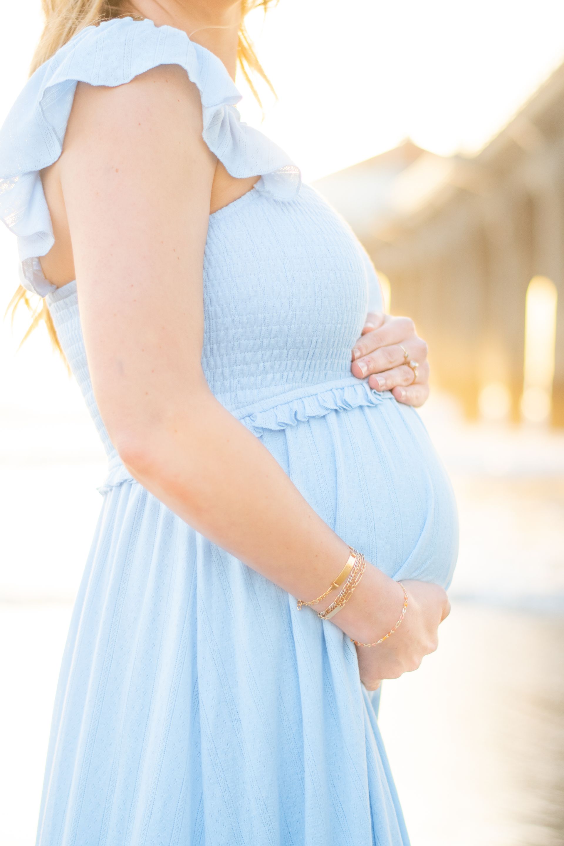 A person in a light blue sundress cradles their pregnant belly while standing outdoors in soft, golden sunlight.