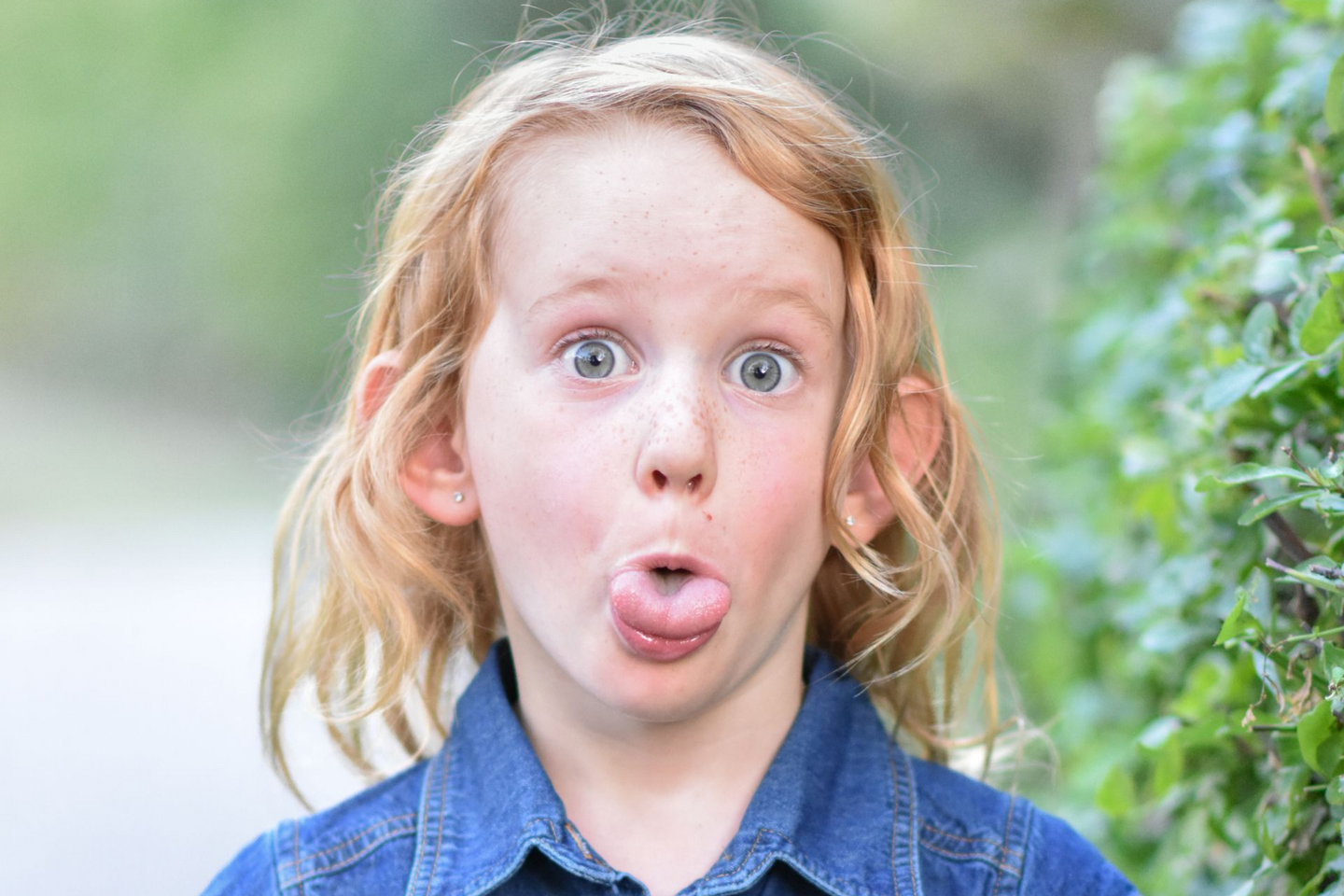 Blond child with wide eyes and tongue sticking out, wearing a blue denim shirt, green foliage background.