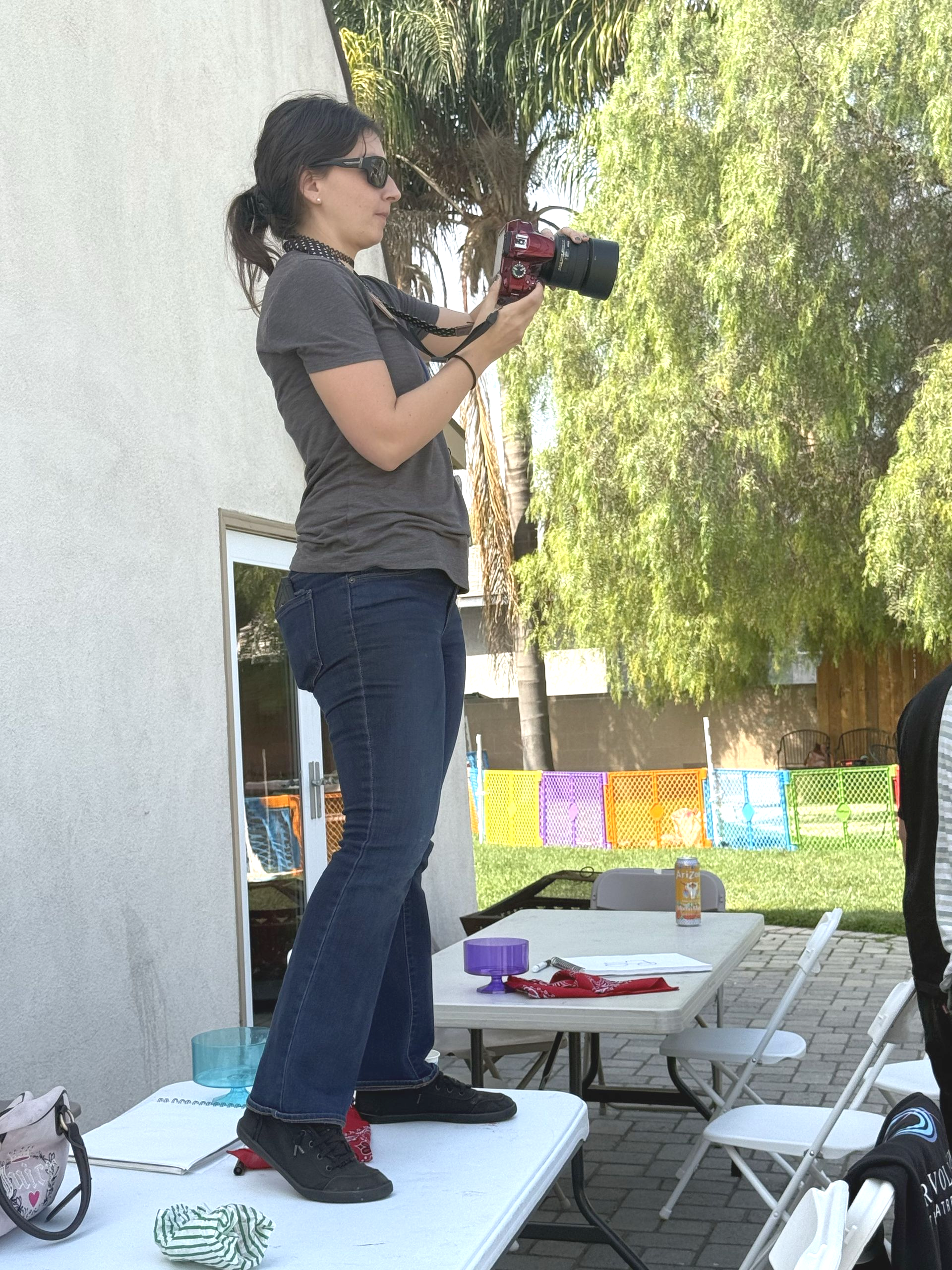 Woman standing on table, holding camera, taking pictures outside.