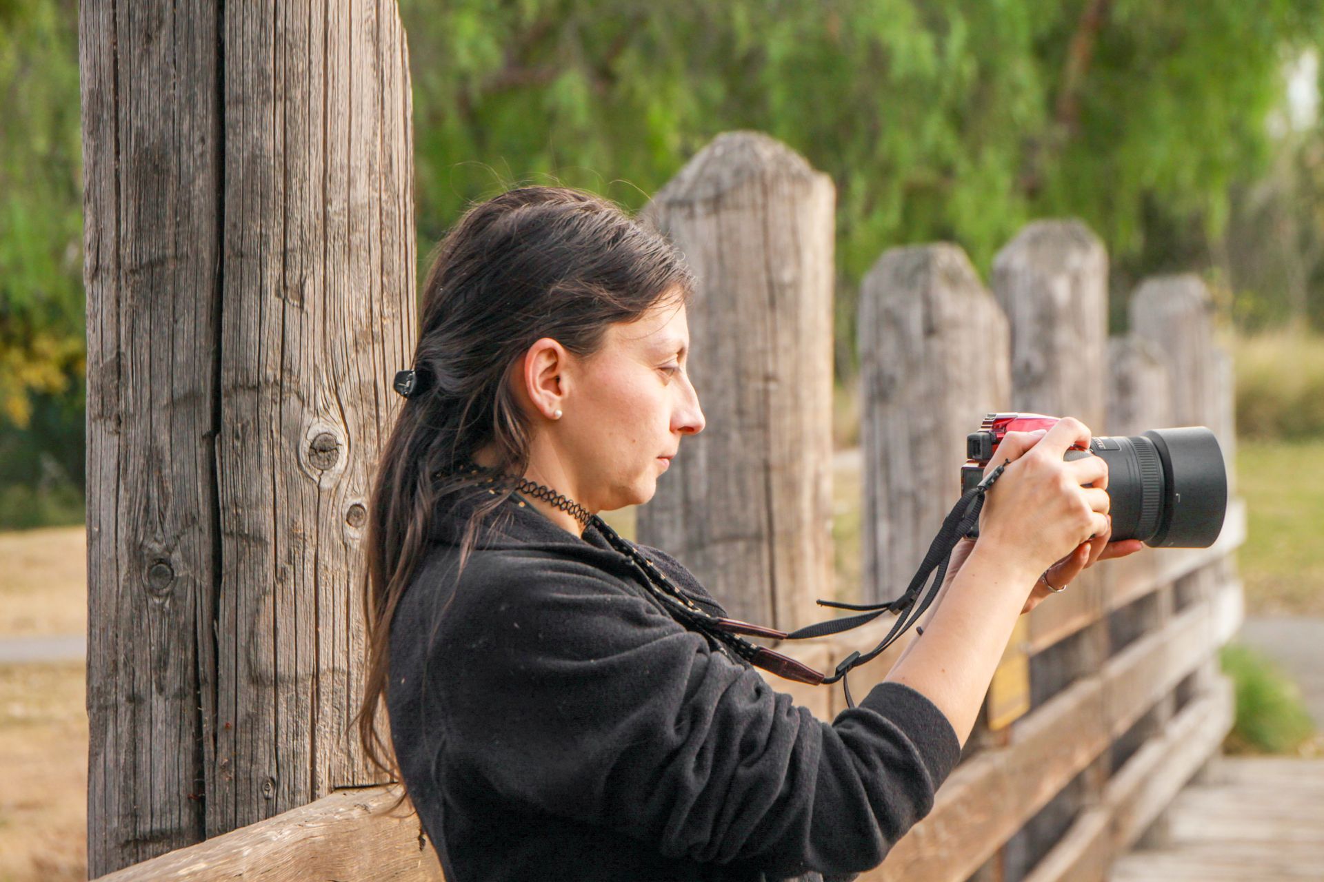 Woman holding a camera, taking a picture outside, standing near wooden posts and a bridge, wearing a black jacket.