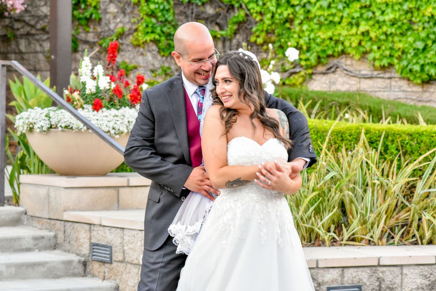 Bride and groom embrace on stairs, floral backdrop. She wears a strapless gown, he a suit. Smiling faces.