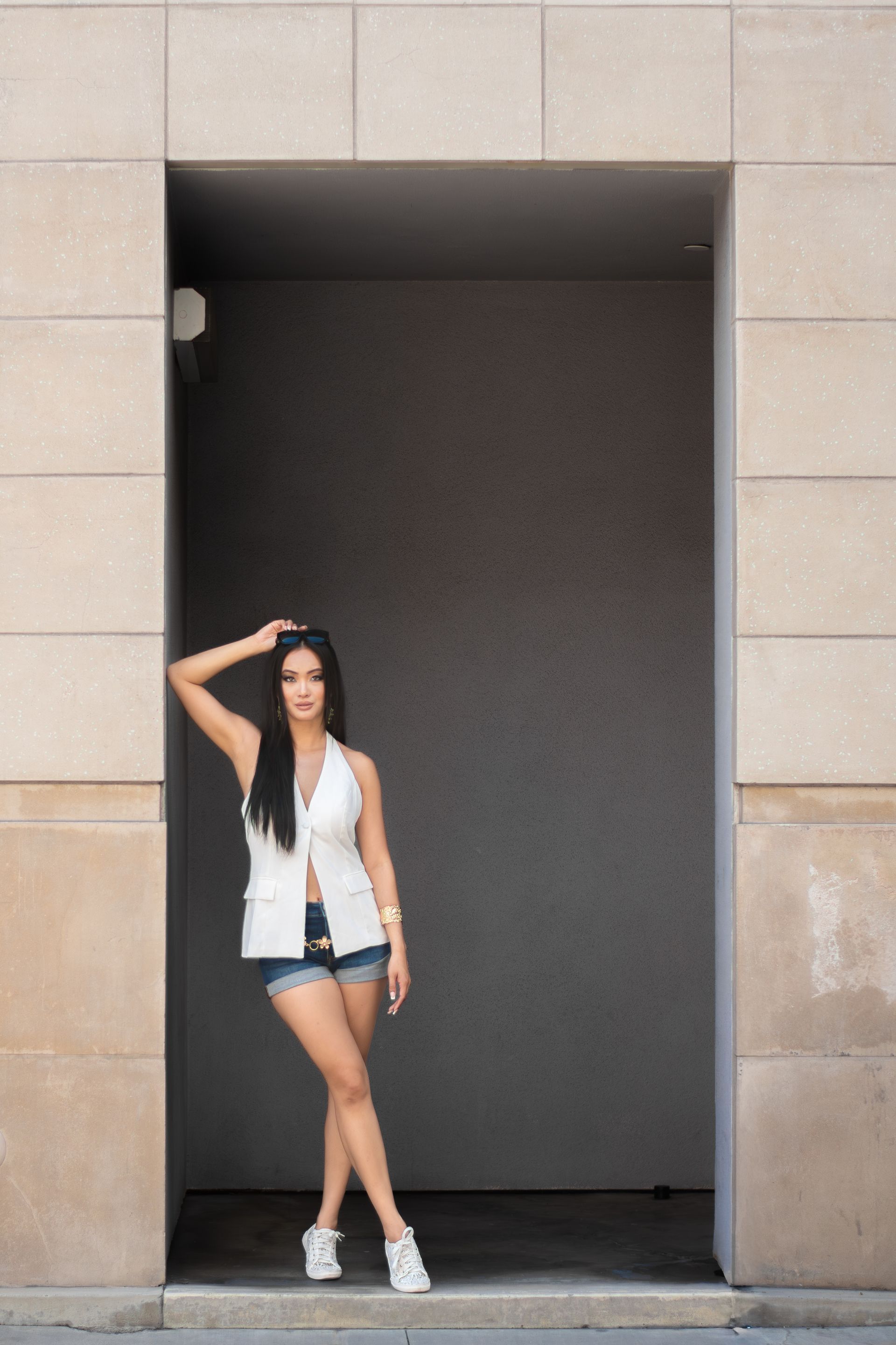 Woman posing in a doorway, wearing a white top, denim shorts, and sneakers.