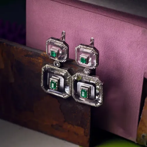 Four silver square earrings with green stones on a pink cushion, beside a dark wooden box. Four silver square earrings with green stones on a pink cushion, beside a dark wooden box.