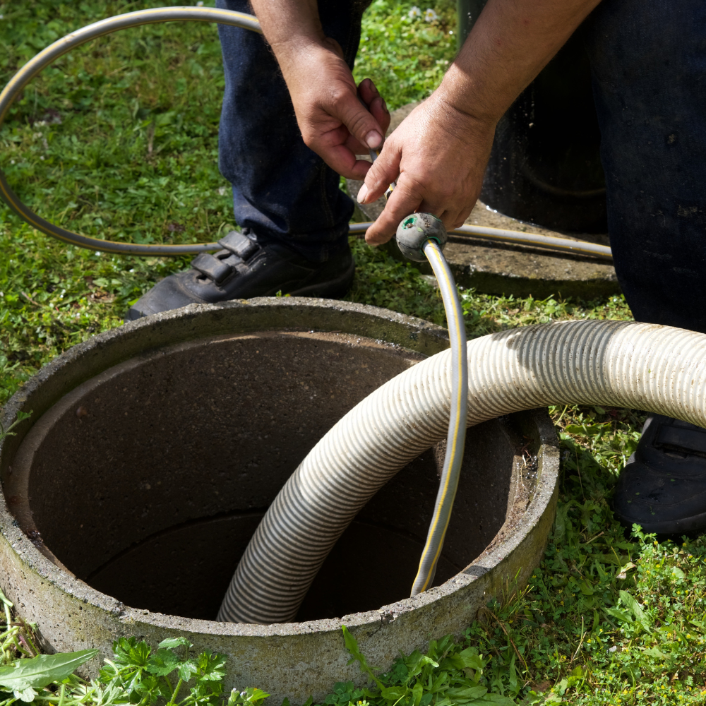A person guides a thick, ribbed suction hose into an open concrete septic tank access port in a grassy area.