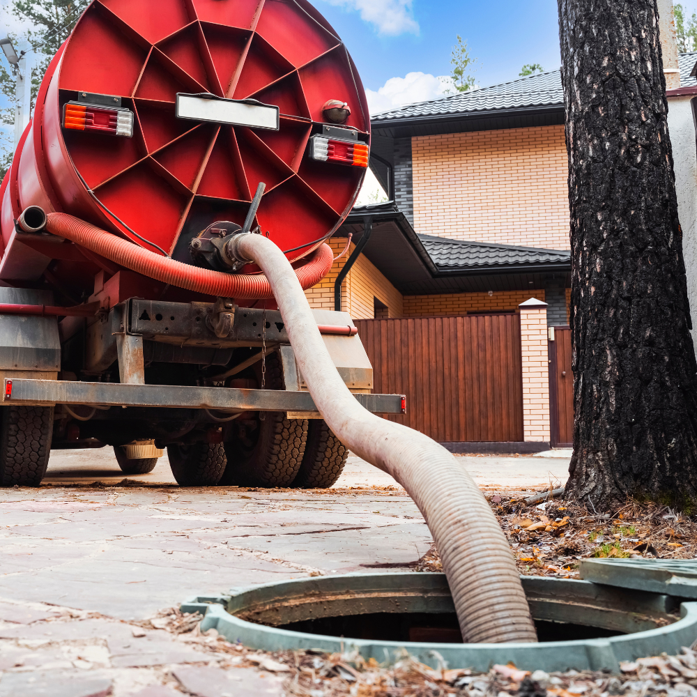 A red vacuum truck hose extends from the rear of the vehicle into an open septic tank manhole near a residential house.