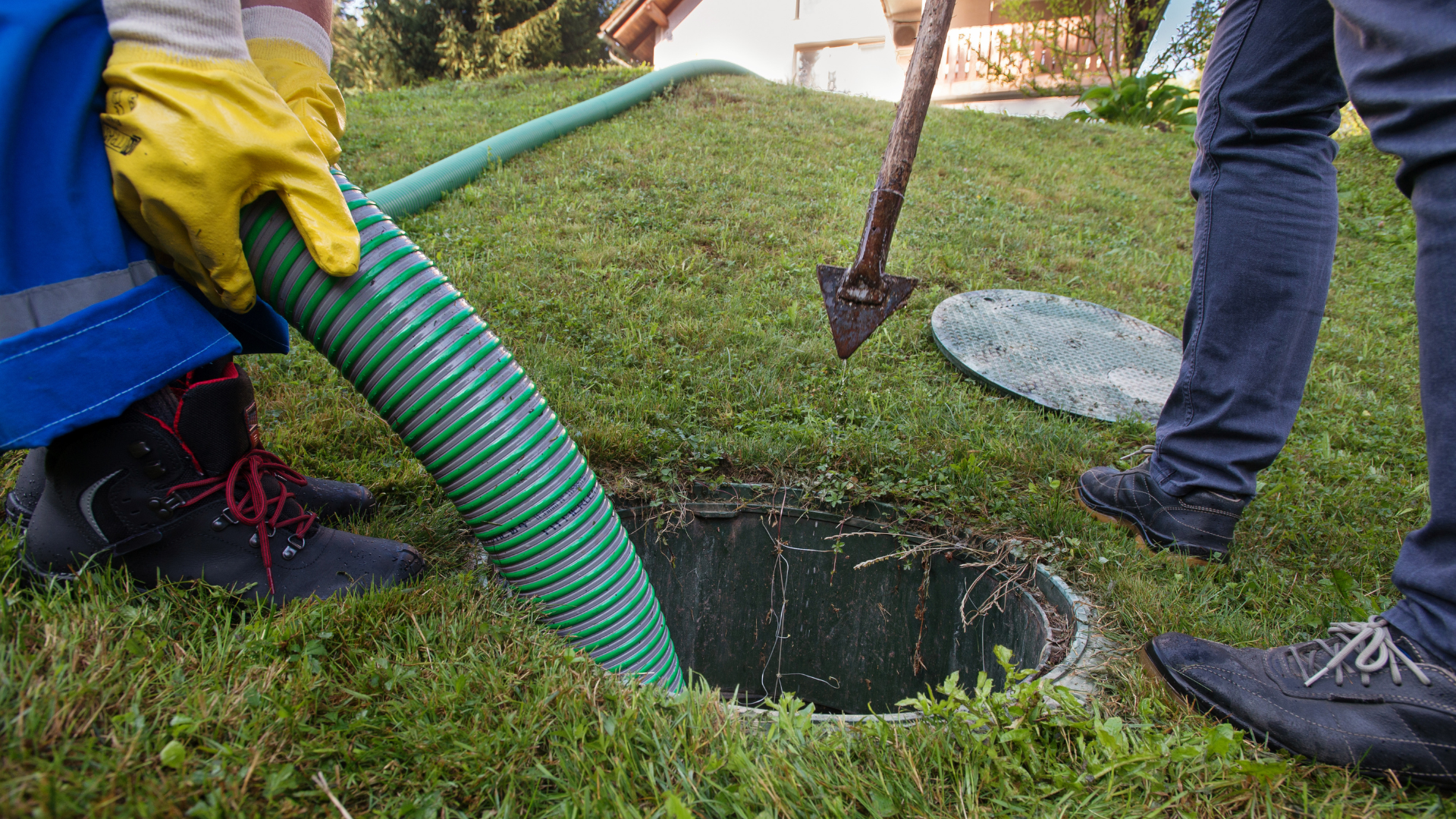 Technicians using a vacuum hose and tool to service an open septic tank in a grassy yard.