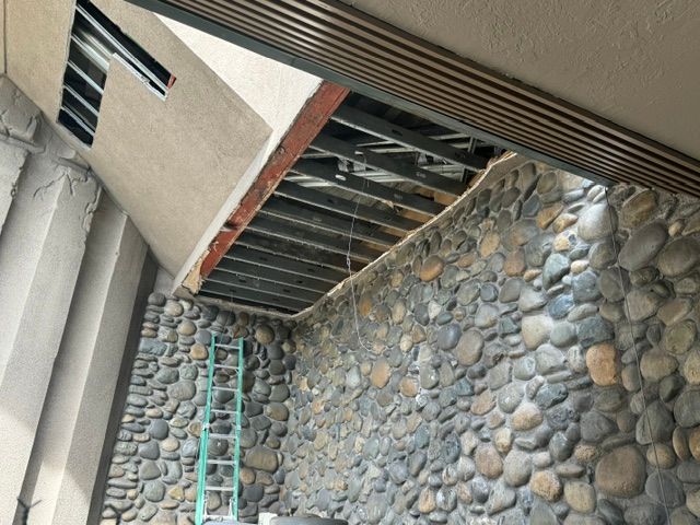 Stone retaining wall under a building overhang with exposed beams and a mint-green ladder.