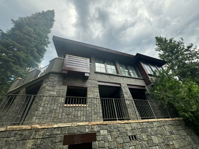 Stone house with dark roof and balconies on a wooded hillside under cloudy sky