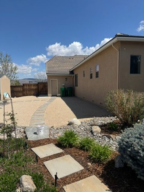 Desert backyard with stepping-stone path, patio, rocks, shrubs, and beige house under blue sky