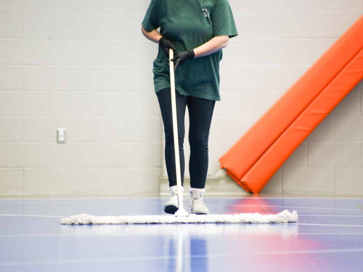 Person in green shirt using a mop on a gym floor beside an orange mat wall
