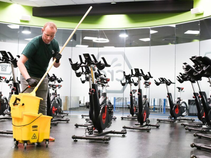 Person cleaning a gym floor with a yellow mop bucket beside stationary bikes