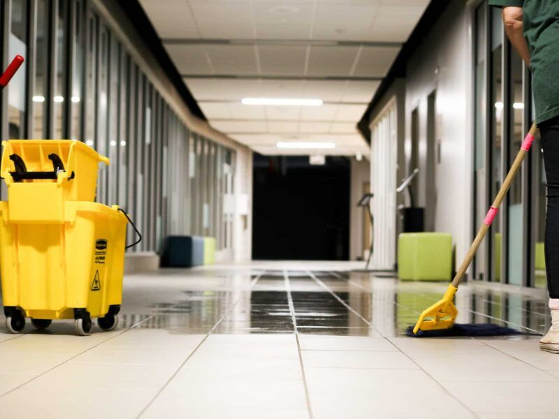 Yellow cleaning cart and mop in a tiled hallway, with a person mopping the floor.