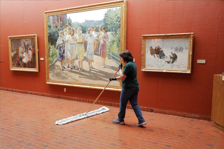 Person mops a red-walled art gallery floor beside framed paintings.