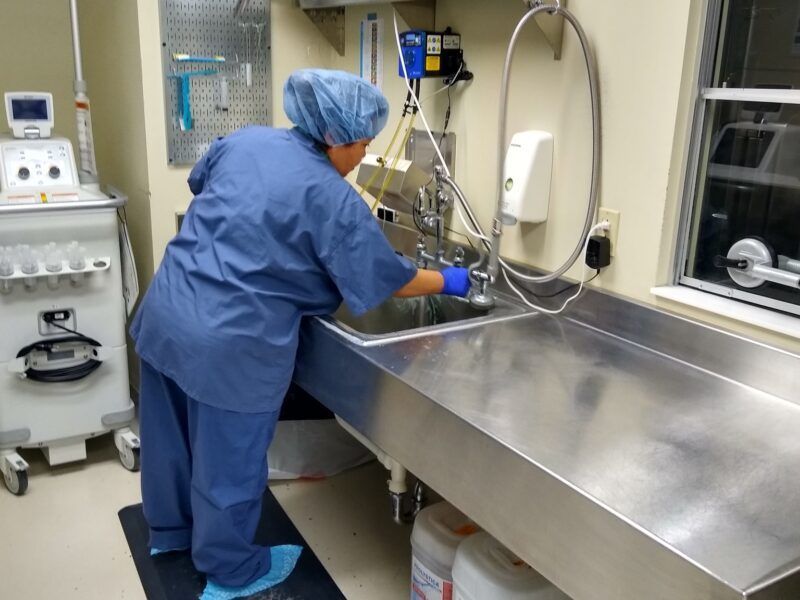 Healthcare worker in blue scrubs washing hands at a stainless steel sink in a clinical room