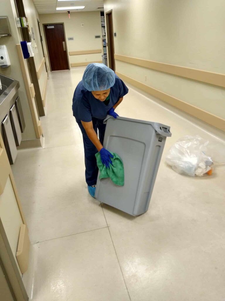Hospital worker in blue scrubs cleaning a hallway with a mop and trash bin
