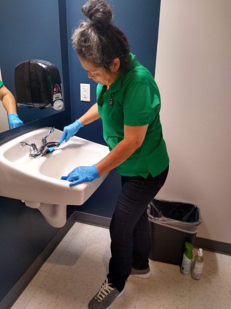 Person cleaning a white sink in a restroom, wearing green shirt and blue gloves.