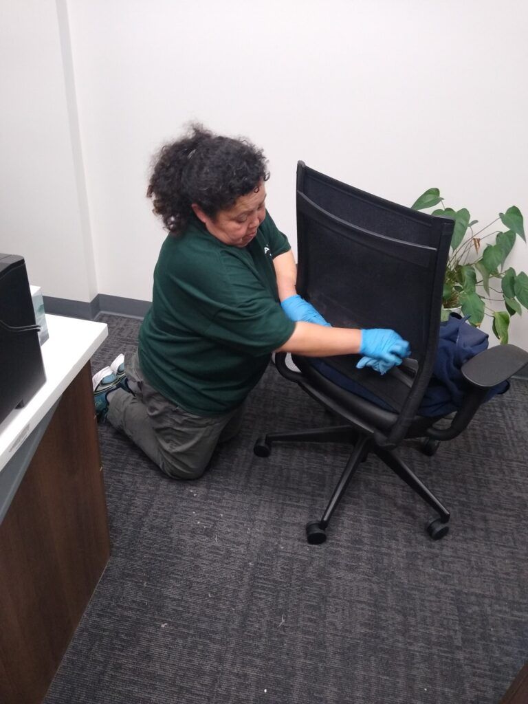Person kneeling beside an office chair, adjusting it in a small room with a desk and plant.