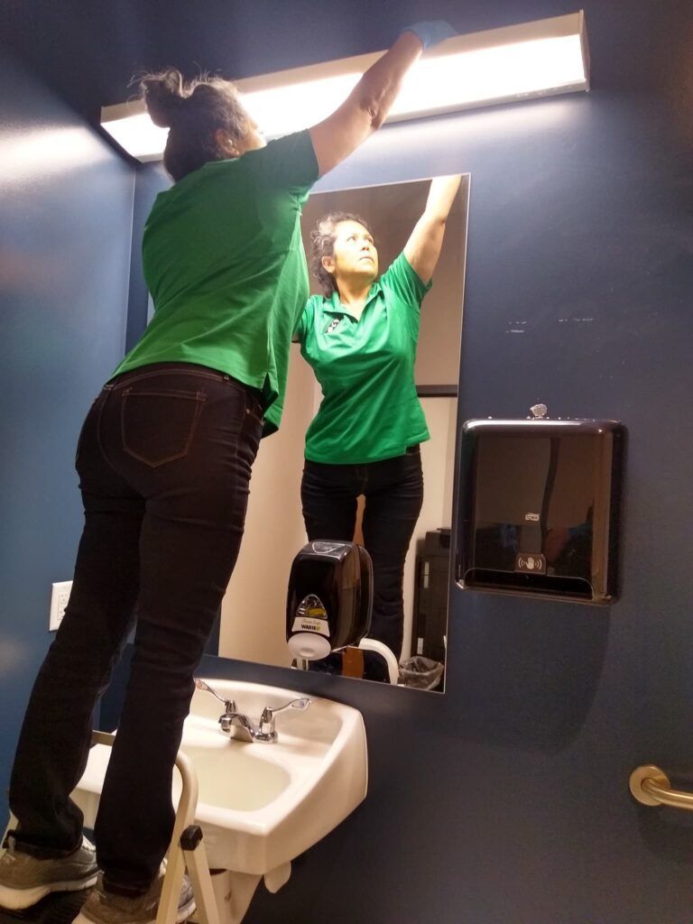 Person in a green shirt cleaning a bathroom mirror above a sink.