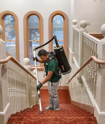 Worker vacuuming a red carpeted stairway in a white-railed hallway